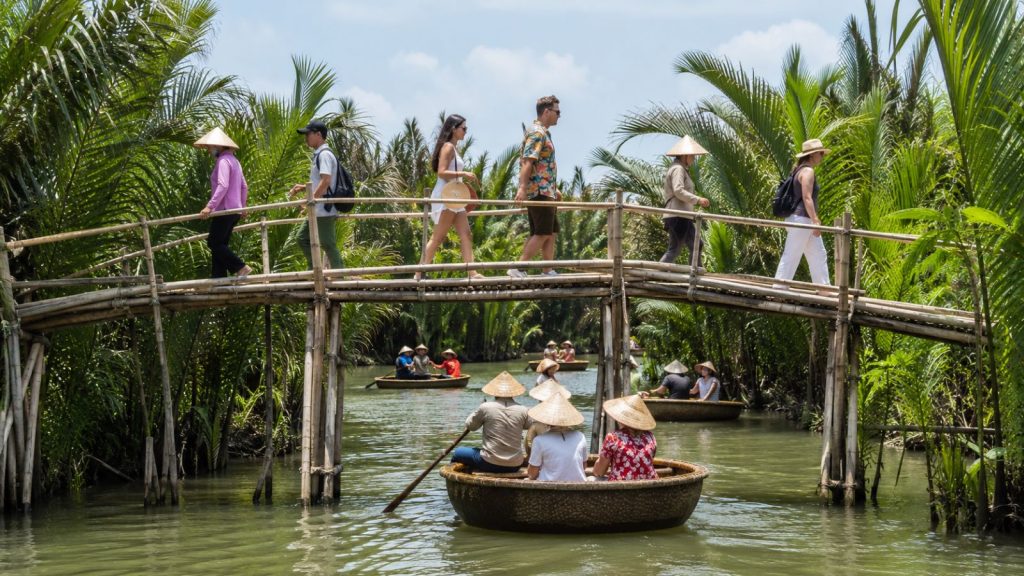 Tourists walking across a bamboo bridge while basket boats pass below through the coconut forest in Hoi An