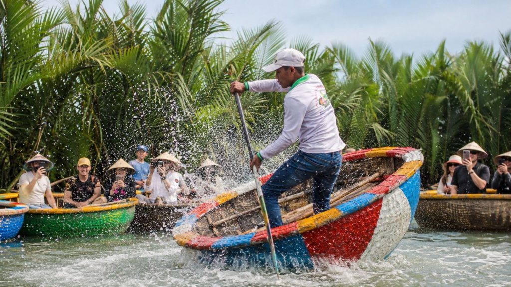 A local boatman performs a spinning basket boat show with splashing water as tourists watch and take photos in Cam Thanh Coconut Forest.