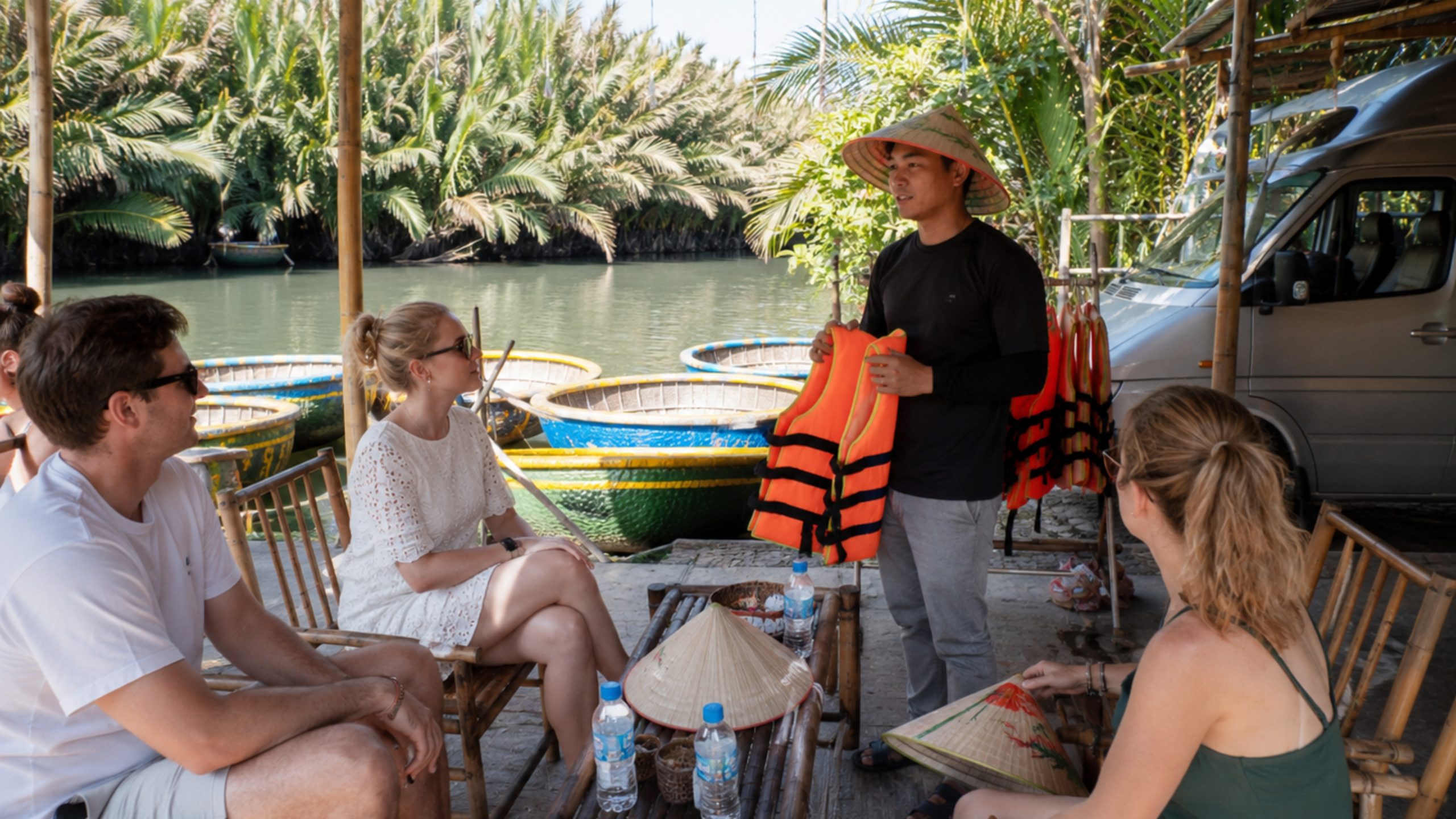 Tour guide preparing visitors for an all-inclusive coconut boat tour with life jackets, bottled water, basket boats, and hotel transfer van nearby.