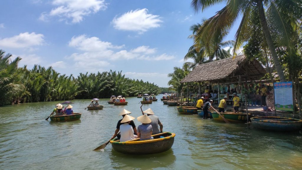 Tourists ride traditional basket boats along the waterway at Bay Mau Coconut Forest in Hoi An, surrounded by lush palms and a riverside hut.