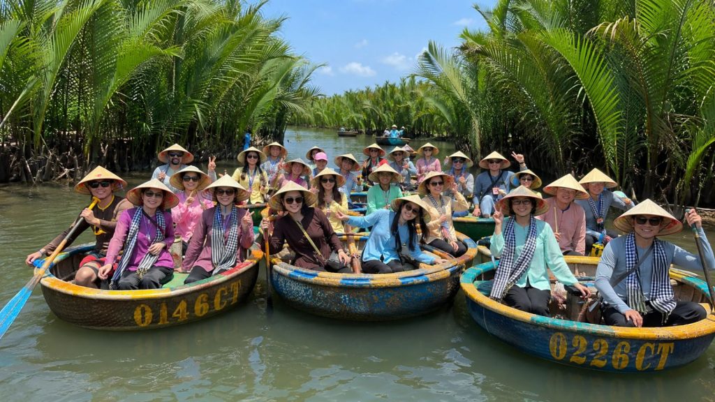 A company group wearing traditional outfits, scarves, and conical hats enjoys a basket boat tour through Cam Thanh Coconut Forest in Hoi An.