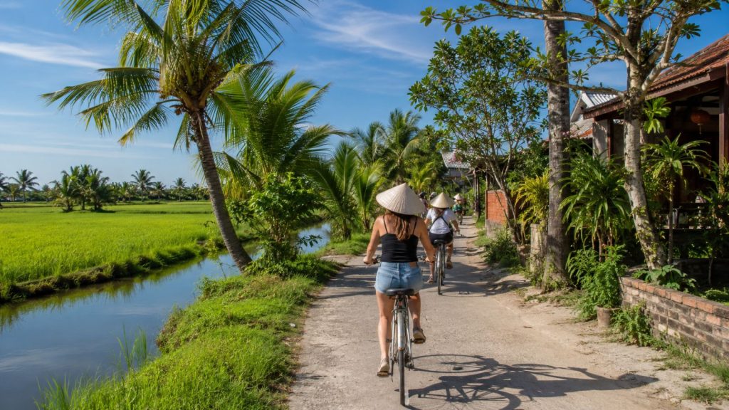 Tourists cycle along a quiet rural path beside rice fields, canals, and tropical greenery in Cam Thanh Village.