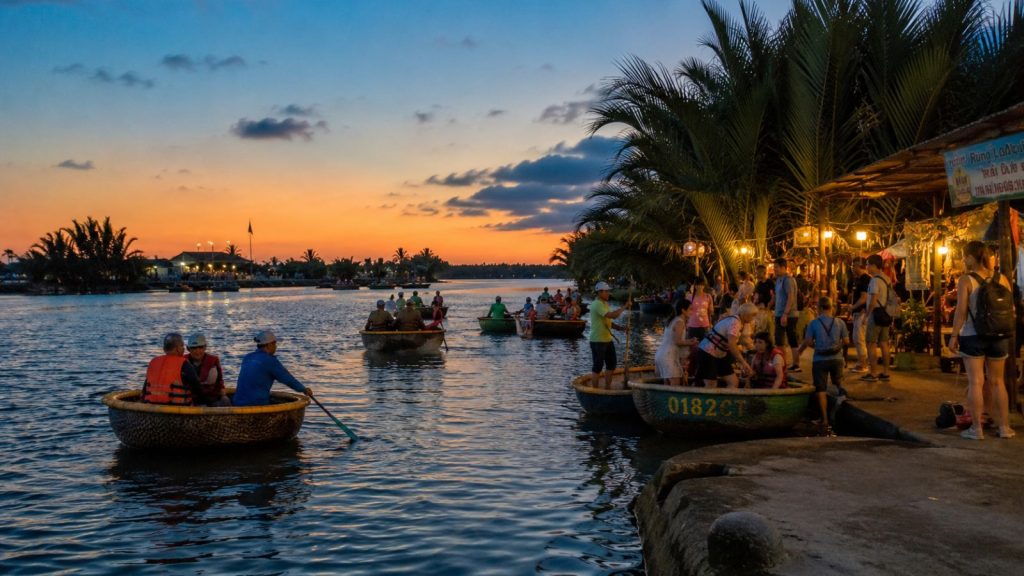 Tourists enjoy basket boat rides at sunset near a lively riverside dock in Hoi An, with warm lights and palm trees along the water.