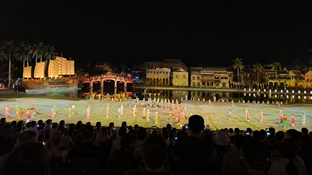 Large outdoor cultural performance at Hoi An Memories Show with actors in traditional costumes, a stage set of old town buildings, and an audience watching at night.