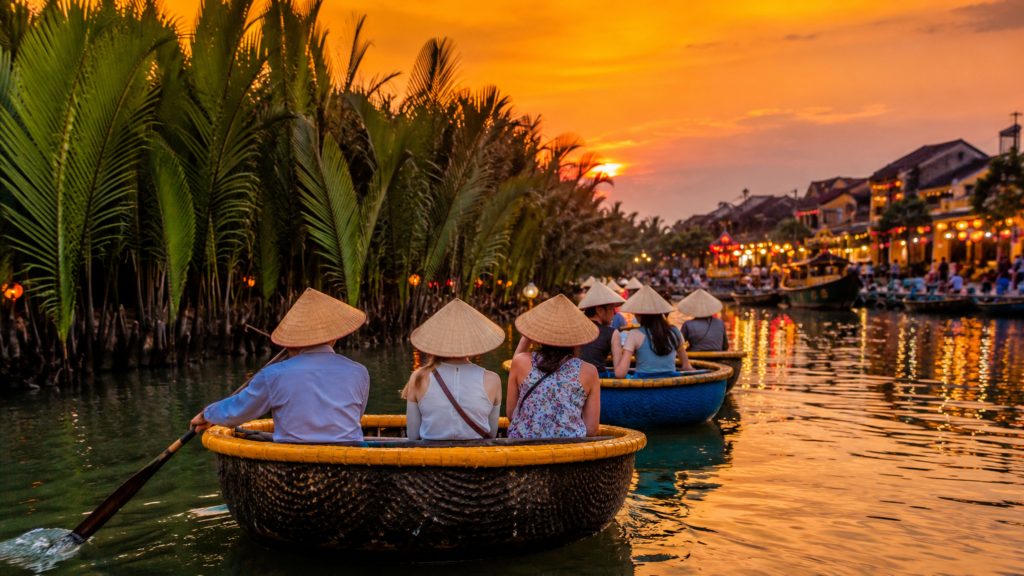 Tourists wearing conical hats riding basket boats along the river at Green Coconut Village during a colorful sunset near Hoi An.