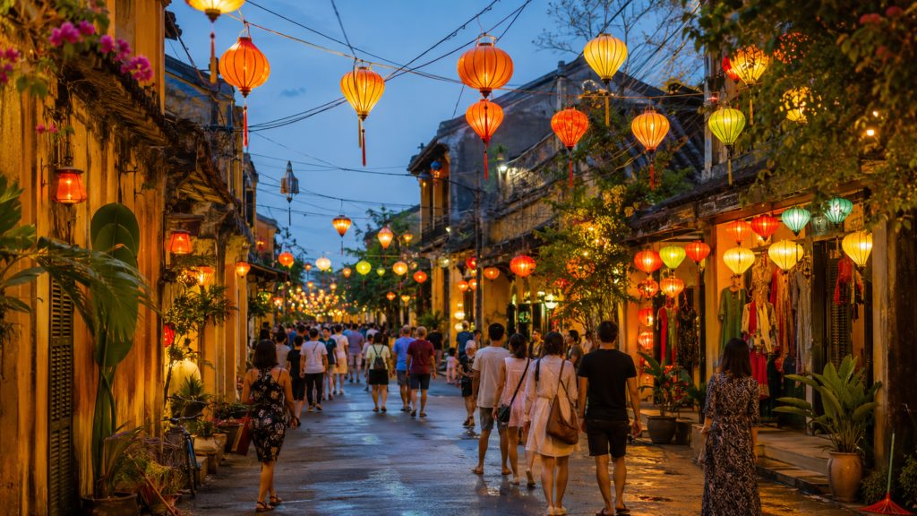 Visitors walking through a lantern-lit street in Hoi An Ancient Town at night, surrounded by traditional yellow buildings, shops, plants, and colorful hanging lanterns.