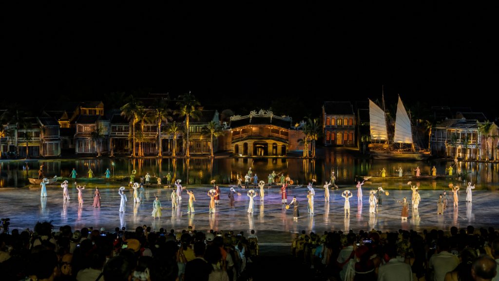 Hoi An Memories Show night performance with actors in traditional costumes on a large outdoor stage beside illuminated old town scenery.