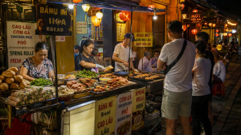 Local vendors preparing Vietnamese street food at a busy Hoi An night market stall, with bánh mì, cao lầu, spring rolls, grilled skewers, and warm lantern lights.