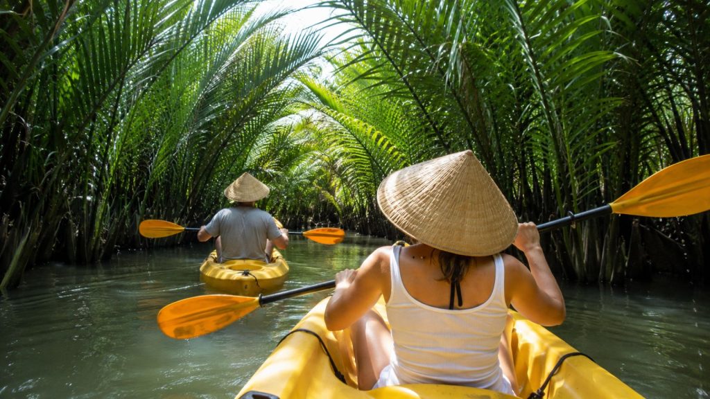 Visitors paddle yellow kayaks through a shaded nipa palm canal in the Cam Thanh coconut forest.
