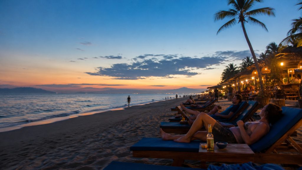 Visitors relaxing on beach loungers at An Bang Beach during sunset, with palm trees, warm beachside lights, gentle waves, and a peaceful evening sky.