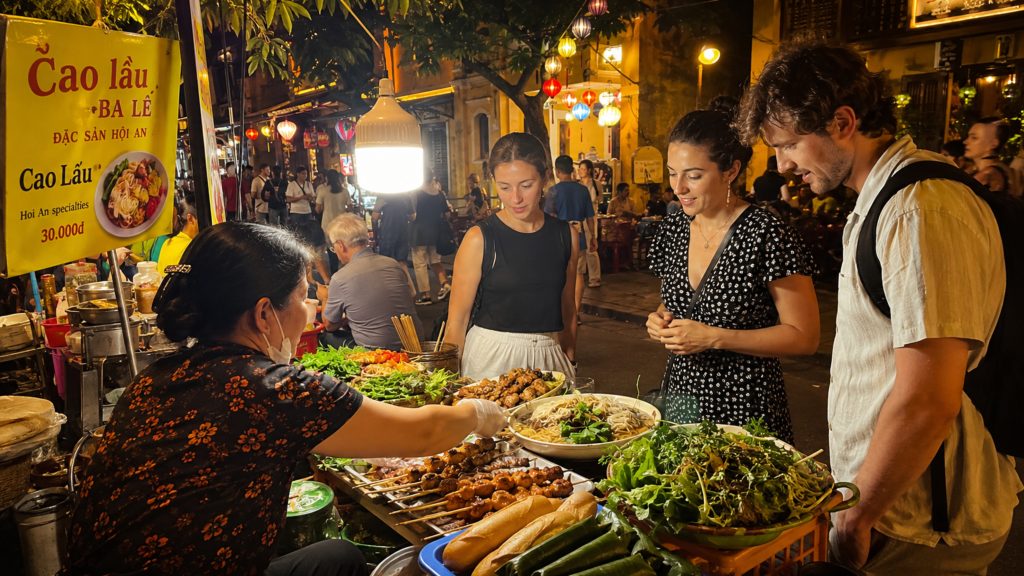Visitors exploring a Hoi An street food stall at night, with a local vendor serving cao lầu, grilled skewers, fresh herbs, noodles, and other Vietnamese dishes under warm lantern lights.