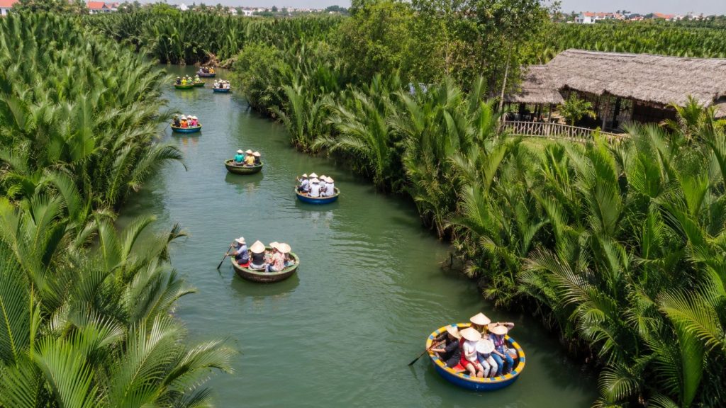 Scenic view of Cam Thanh Coconut Forest with basket boats traveling along a green waterway beside a rustic thatched riverside hut.