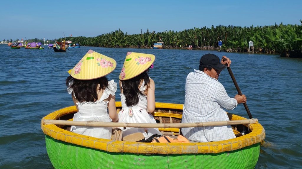 Two tourists wearing conical hats sitting in a green and yellow basket boat while a local guide paddles on the river
