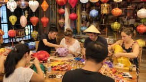 Travelers learning to craft traditional Vietnamese lanterns in a Hoi An workshop filled with colorful hanging lanterns.