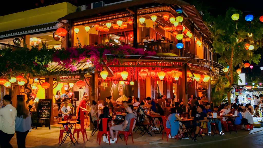 Guests relaxing at a vibrant Hoi An bar and pub at night, surrounded by colorful lanterns, outdoor seating, warm lights, and a lively evening street atmosphere.